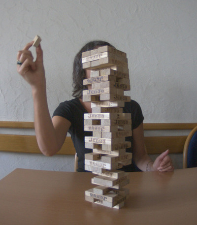 Eine Frau legt einen Stein auf einen Jenga-Turm. Der Turm ist bereits sehr hoch gebaut.